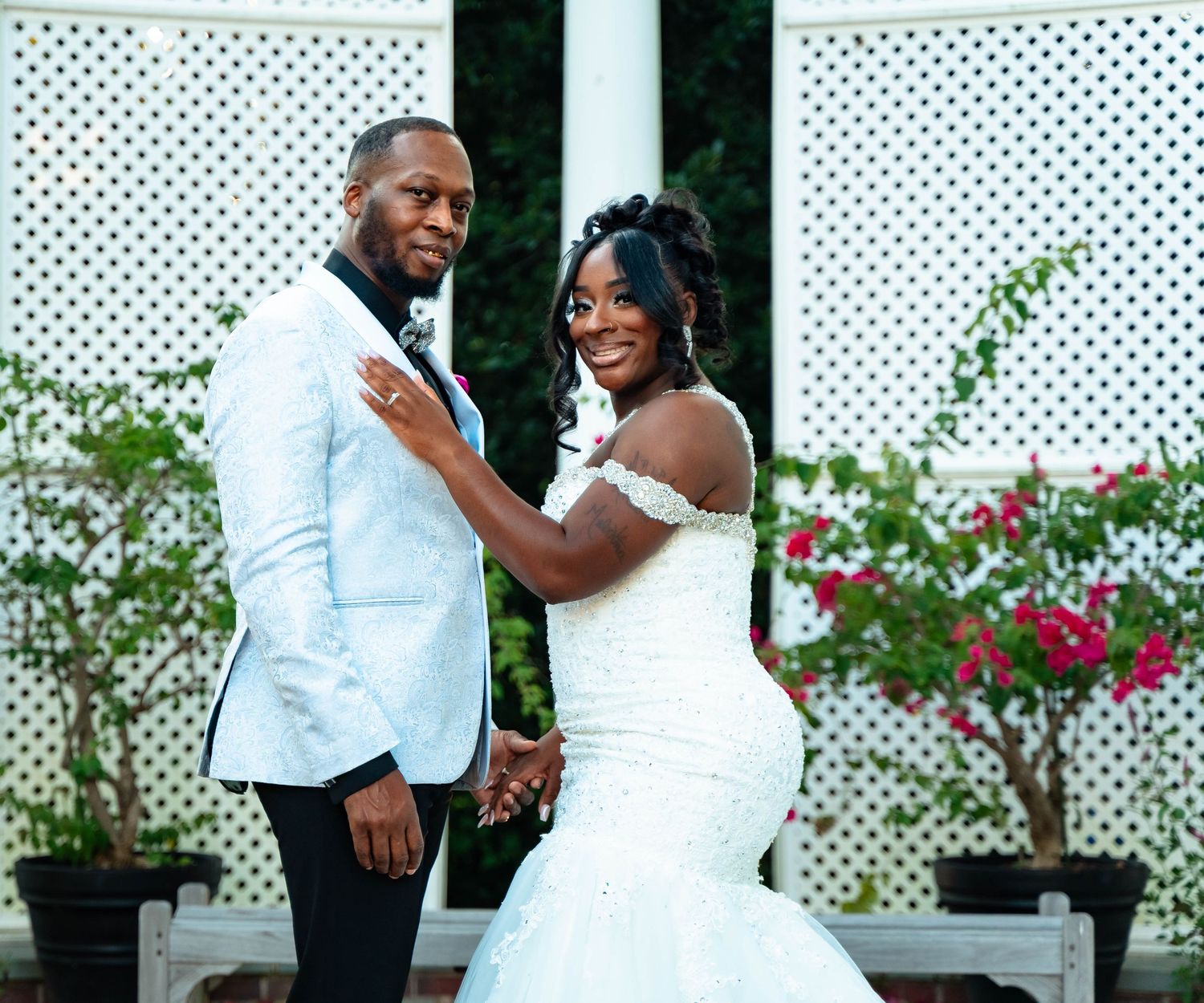 Newlyweds pose together against white lattice backdrop with red flowers.