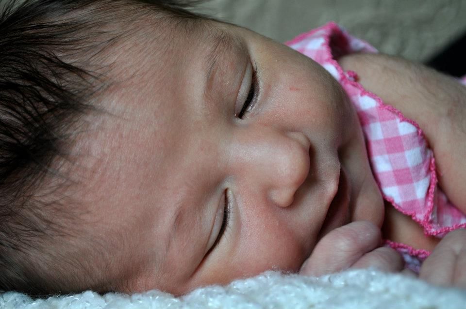 Sleeping newborn baby wearing pink gingham clothing on white blanket.