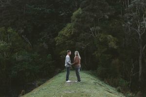 A couple stands facing each other on a grassy path surrounded by lush forest.