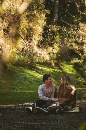 A couple shares an intimate moment sitting together on the ground in a sunlit forest setting.