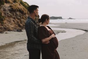 An expectant couple stands together on a dramatic coastal beach at sunset.