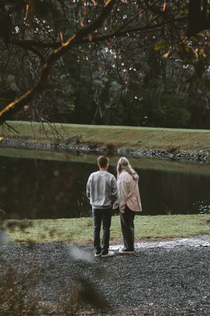 Two figures stand together looking out over a serene lake surrounded by trees.