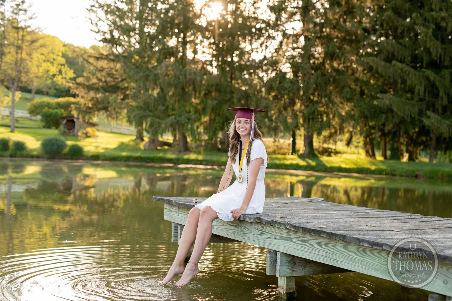 Graduate in white dress sits on wooden dock dangling feet over tranquil pond.