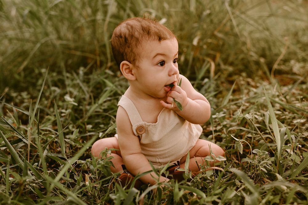Baby in tan romper sitting in grass with finger in mouth during outdoor photoshoot.