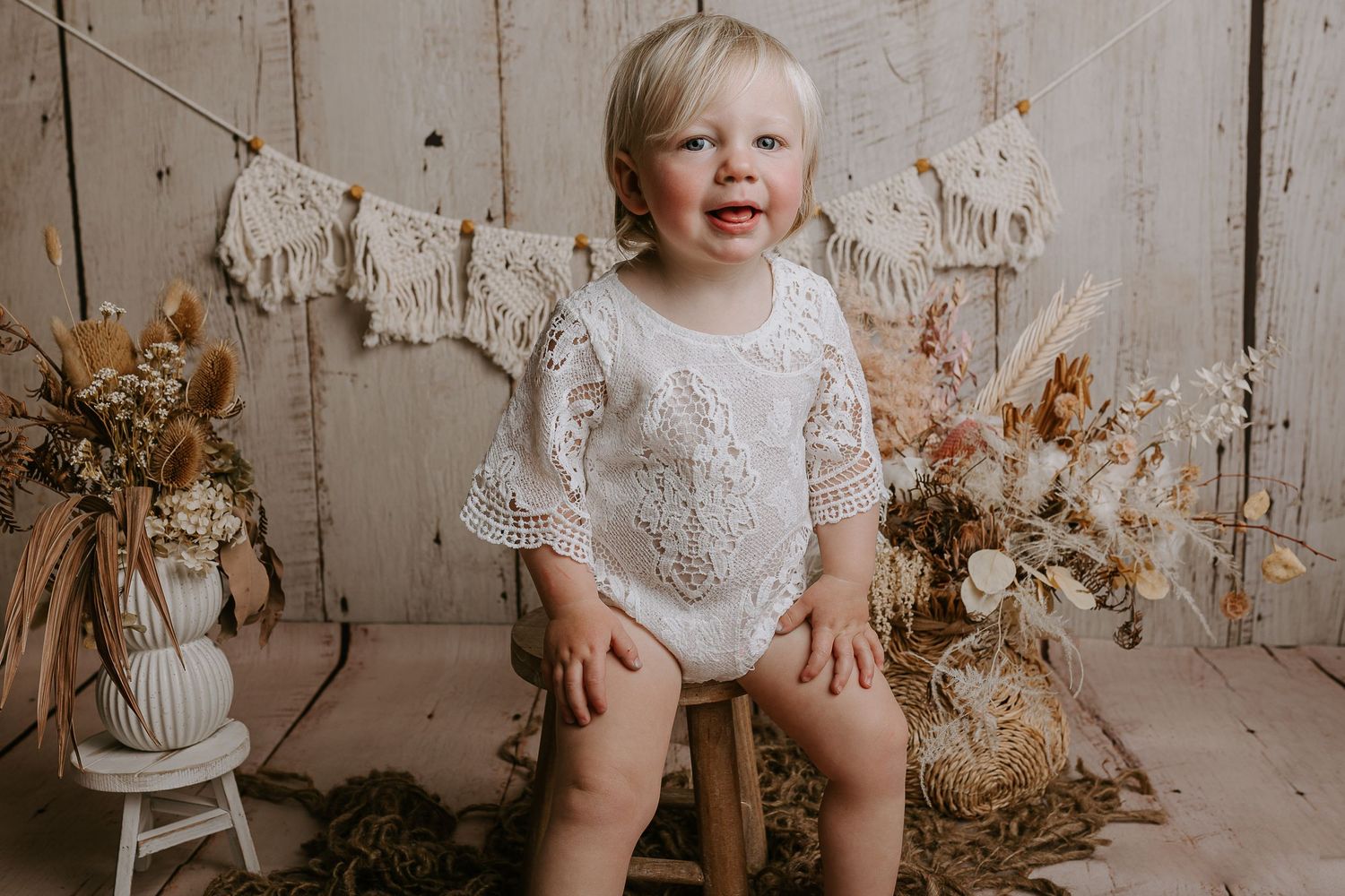 Baby wearing white lace outfit poses in wicker basket with dried floral decorations.