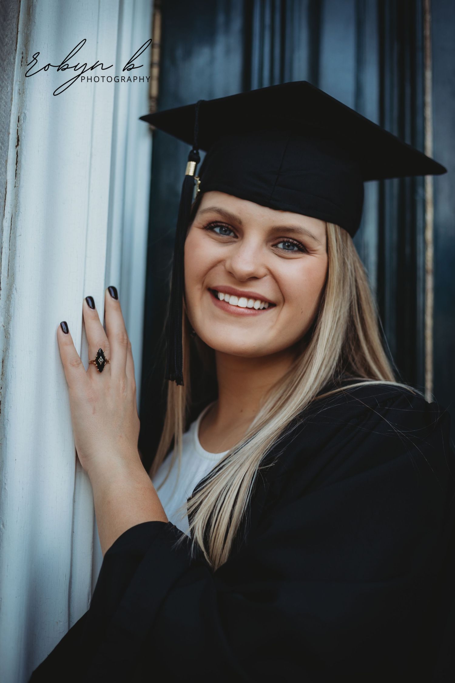 A graduate in traditional black cap and gown poses with a bright smile against a curtained backdrop.