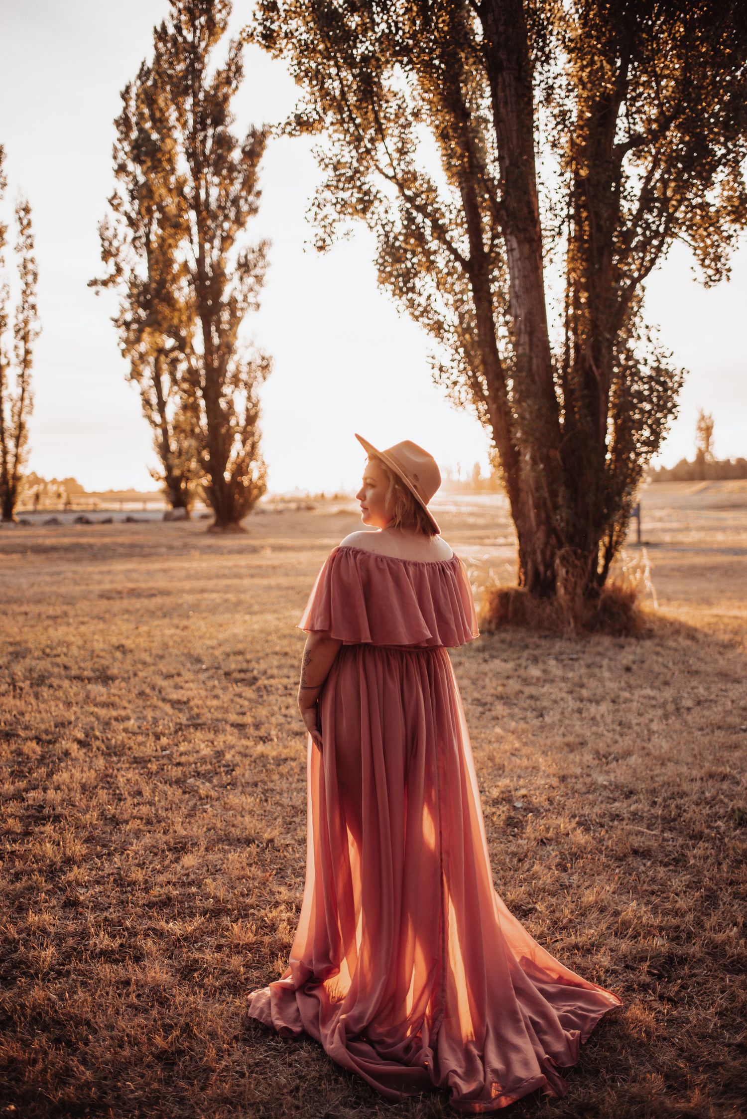 Person in flowing coral dress and hat standing near tall trees at sunset.
