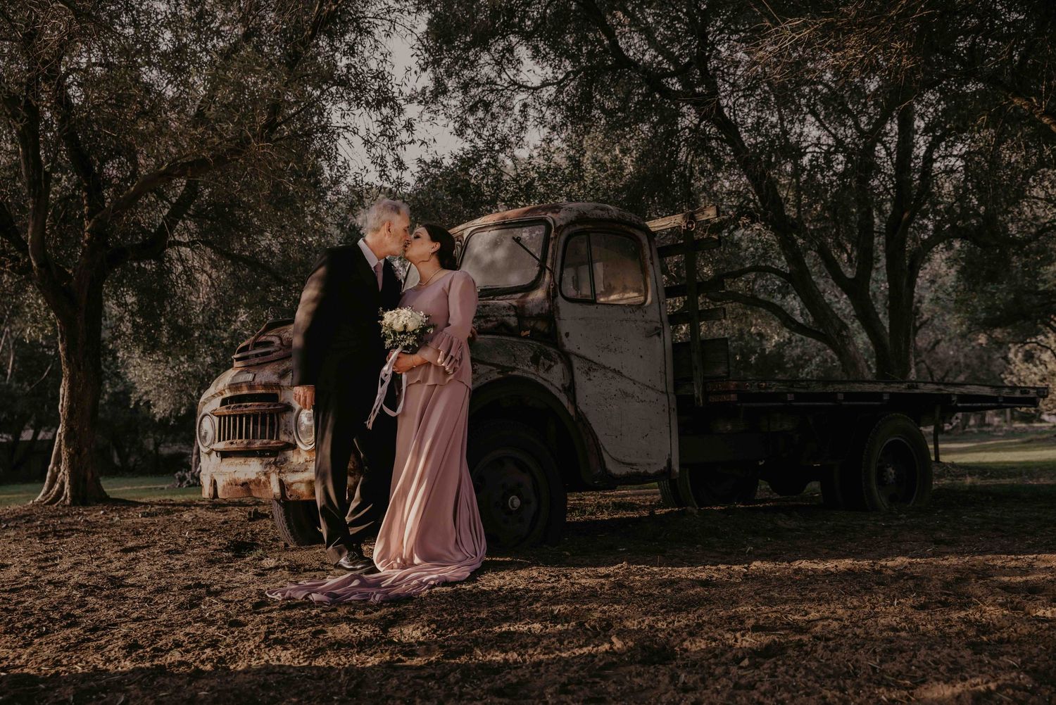 Couple in elegant attire dancing near vintage truck at sunset with trees in background.