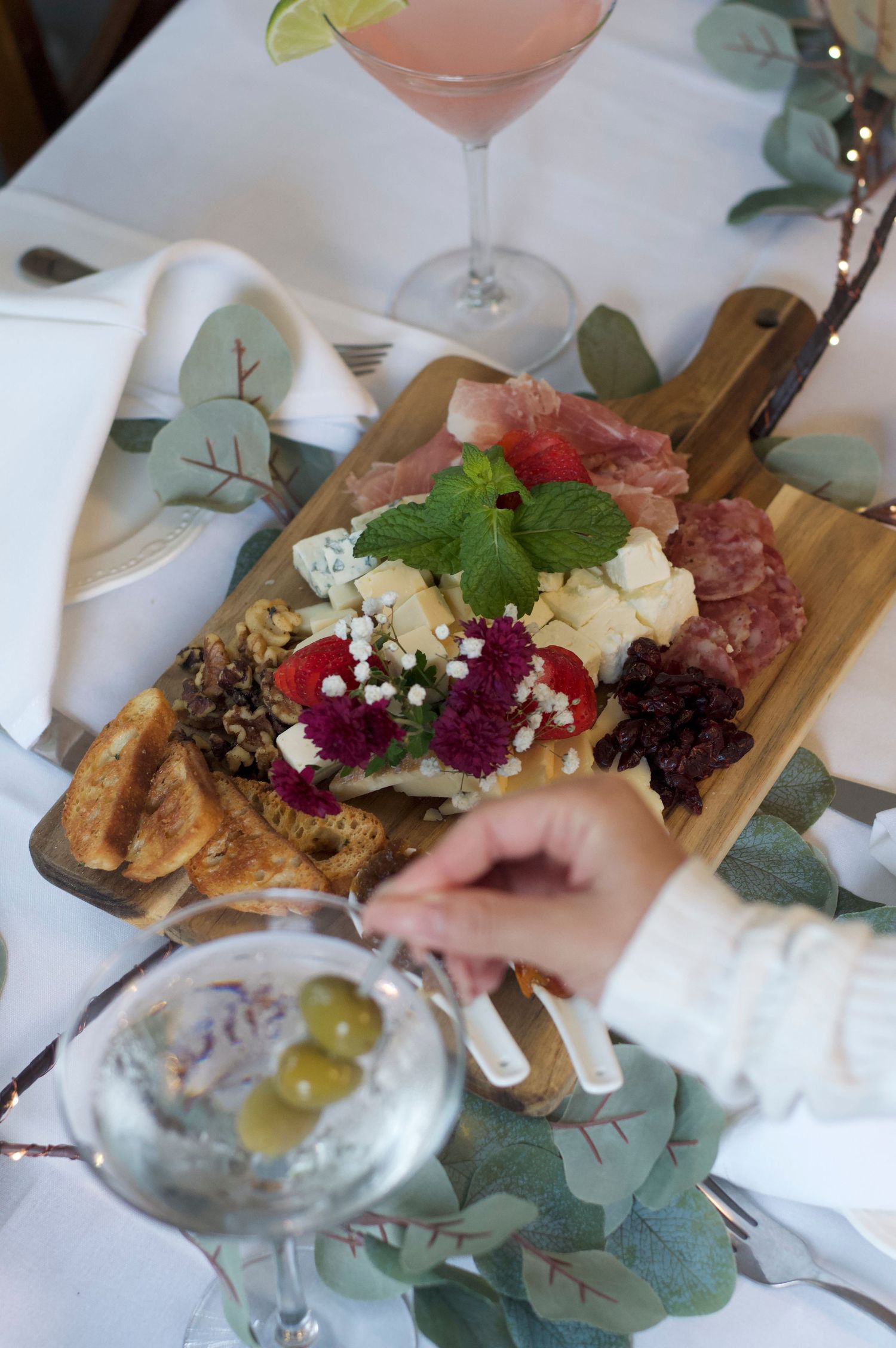 An elegant charcuterie board with meats, cheeses, and garnishes served alongside cocktails on a white tablecloth.