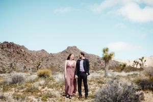 A couple stands together against a desert landscape with mountains and joshua trees.