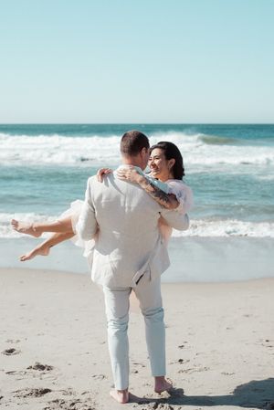 Playful couple dancing together on beach in white outfits.