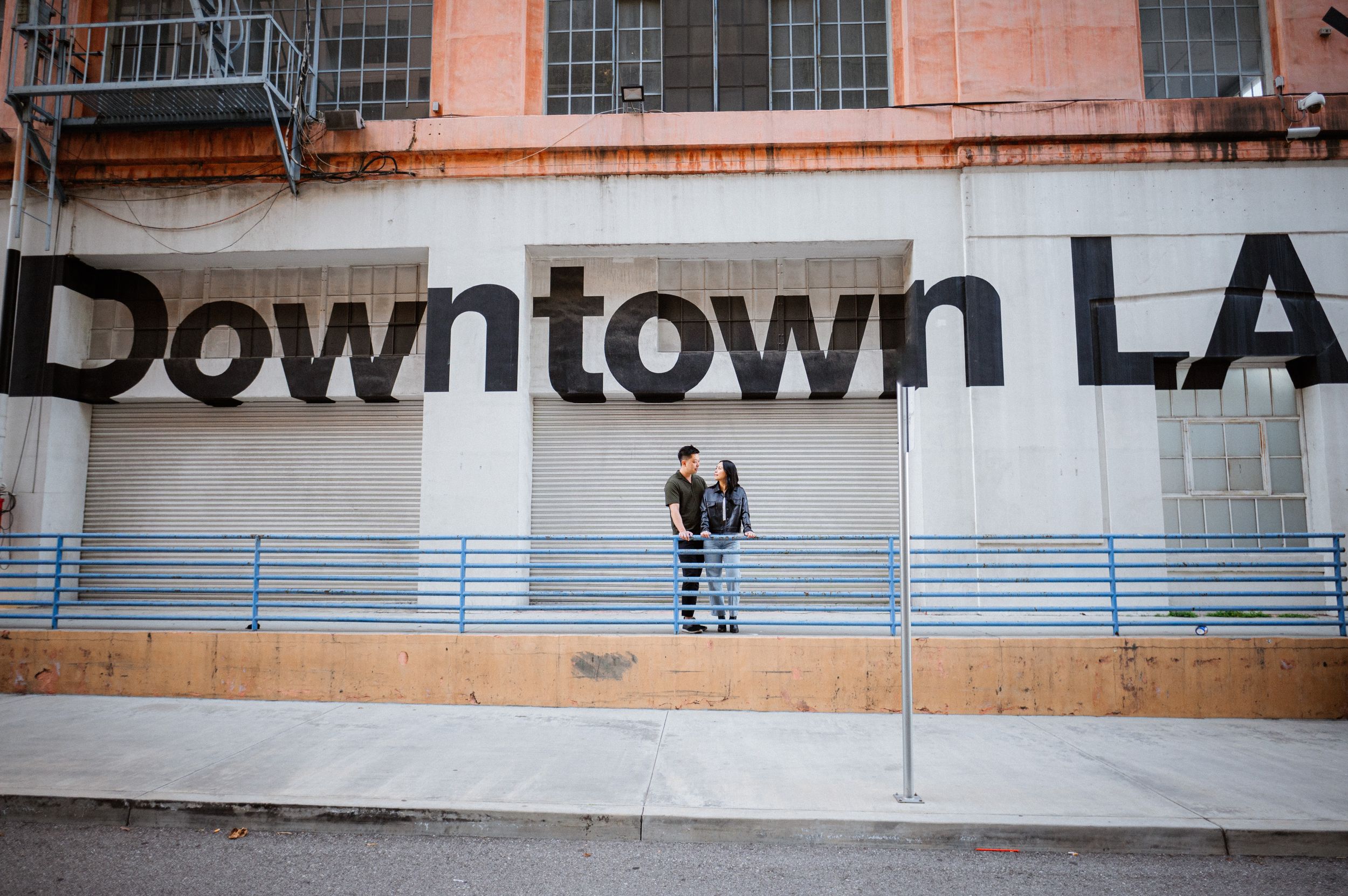 A couple poses in front of a Downtown LA sign on an industrial building wall.