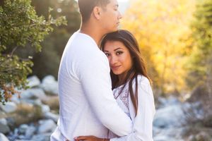 A couple in white outfits embraces during a romantic outdoor photoshoot with golden sunlight filtering through trees.