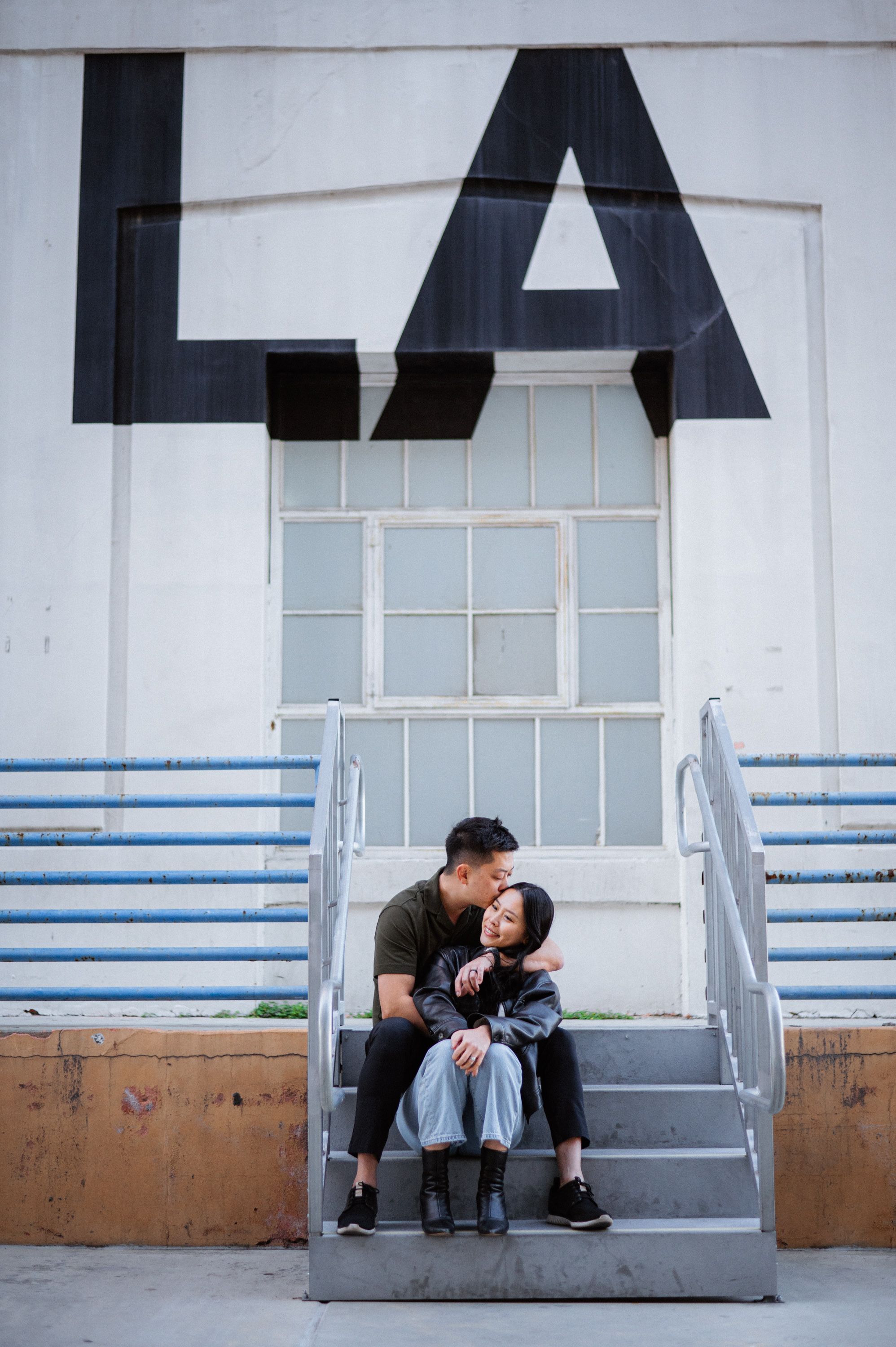 A couple shares an intimate moment sitting on gray steps beneath a large letter A on a white building.