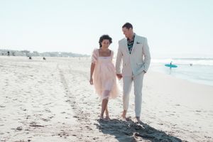 Couple in light colored formal wear walking along sandy beach.