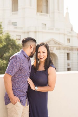 A couple in coordinated navy and purple outfits poses together against a white building.