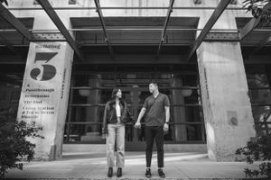 A couple stands together outside a modern building entrance with number 5.