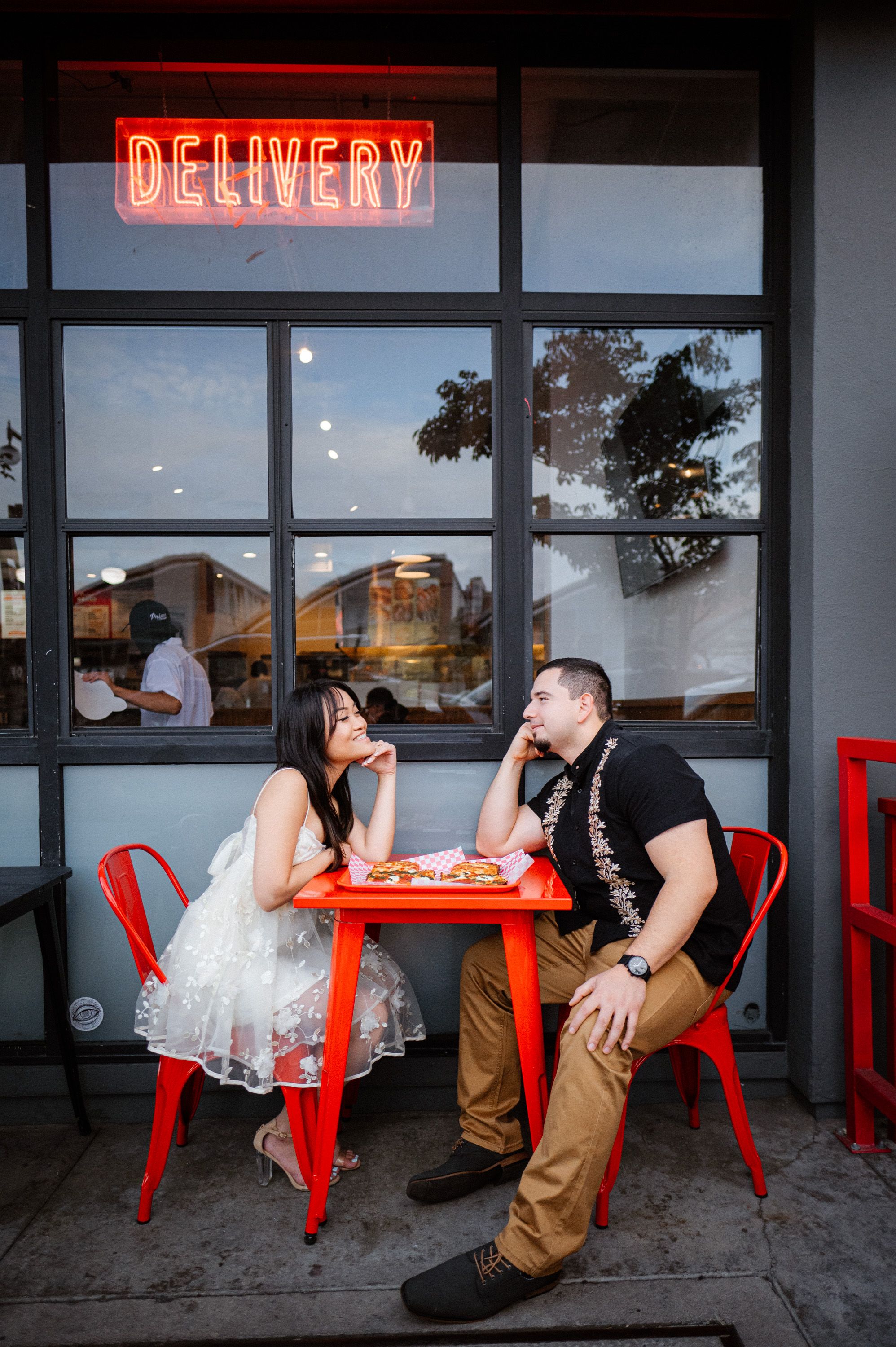 A couple enjoys pizza at a red table under a neon delivery sign.