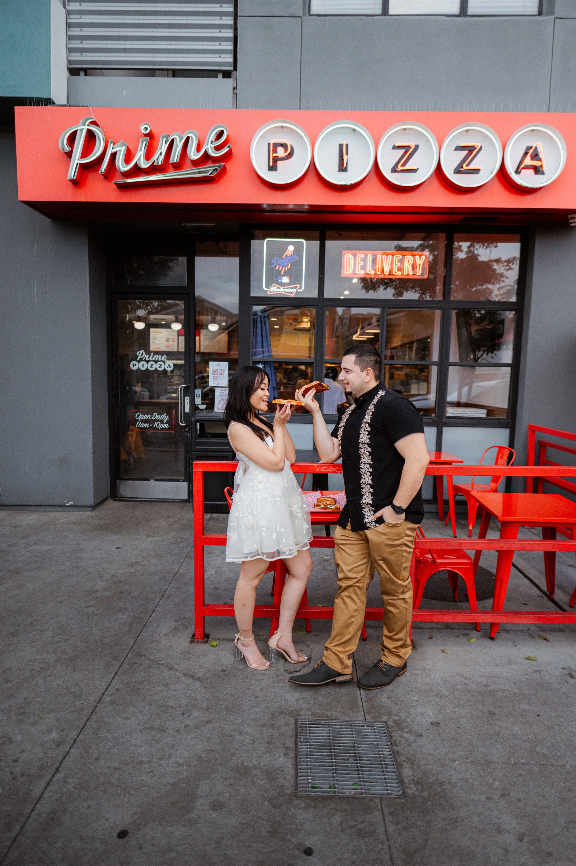 A couple poses outside Prime Pizza restaurant with red signage and chairs.