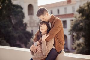 A couple embraces on a balcony with Mediterranean architecture in soft lighting.