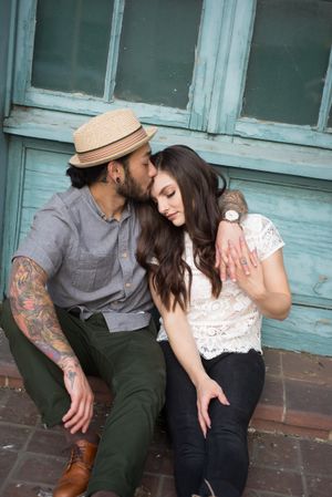 A couple in casual attire sits close together against a weathered blue building exterior.