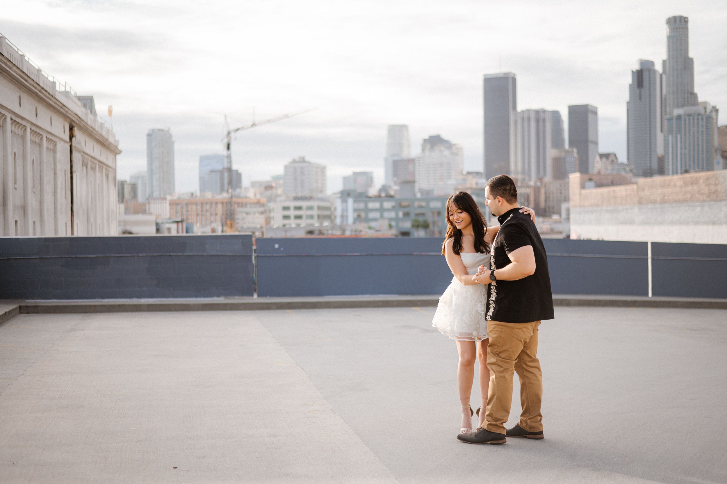 A couple stands together on an urban rooftop with downtown skyscrapers in view.
