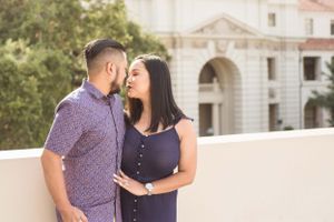 A couple embraces on a balcony with classical architecture in the background.
