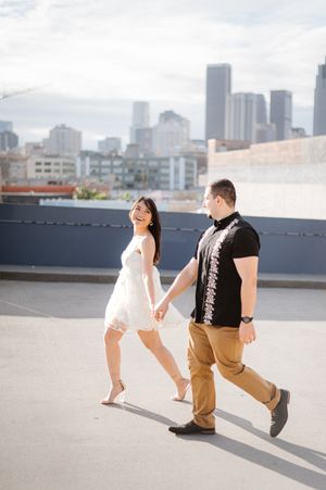 A couple walks hand in hand on a rooftop with city skyline in background.