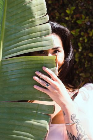 Woman in white dress peeks through large tropical leaf showing tattoo and engagement ring.