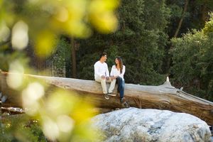 Two people sit together on a large rock formation in a natural outdoor setting surrounded by greenery.