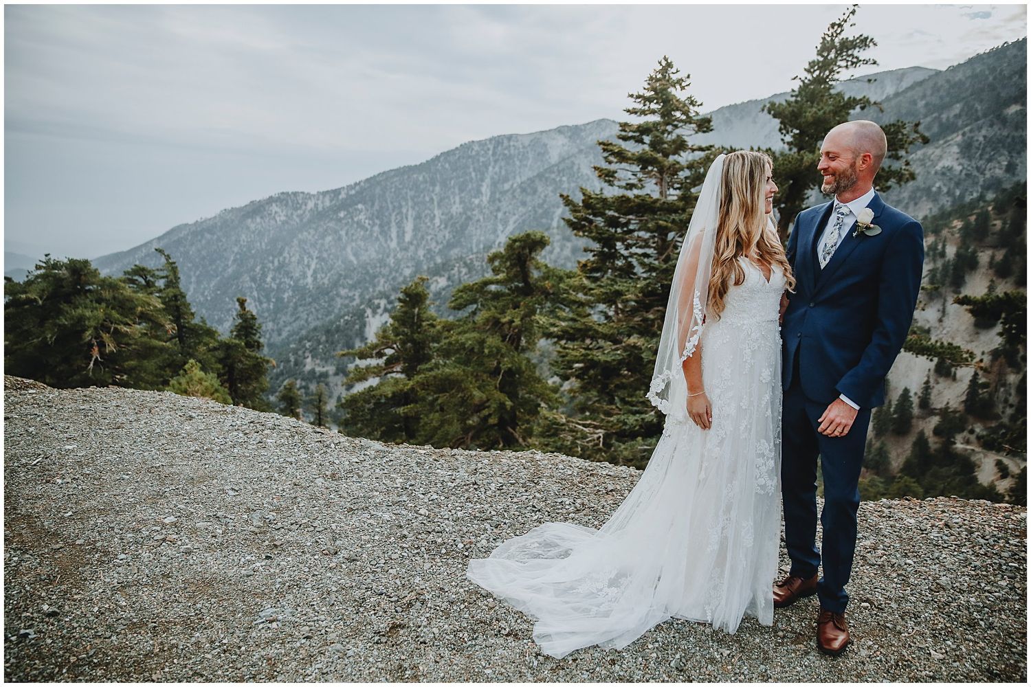 A couple shares a romantic moment on a mountaintop with sweeping views of evergreen trees and rugged peaks in the background.