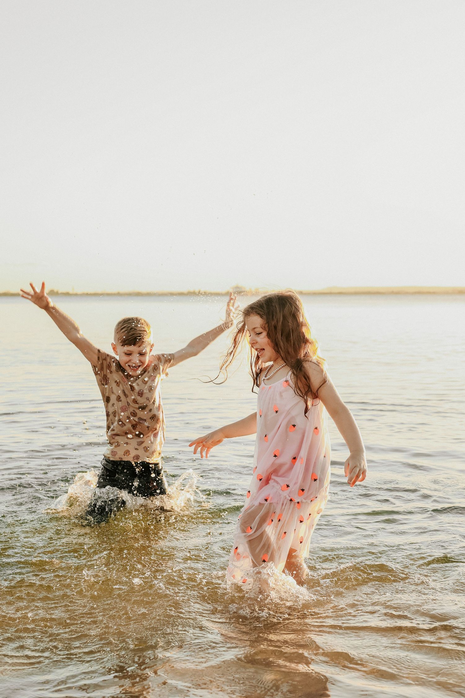 Two children laughing and splashing in shallow ocean water during a fun beach photo session.