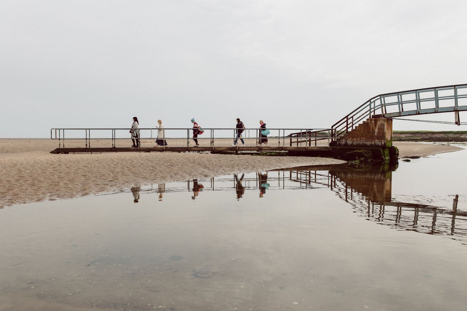 People walking on wooden boardwalk along sandy beach in overcast weather.