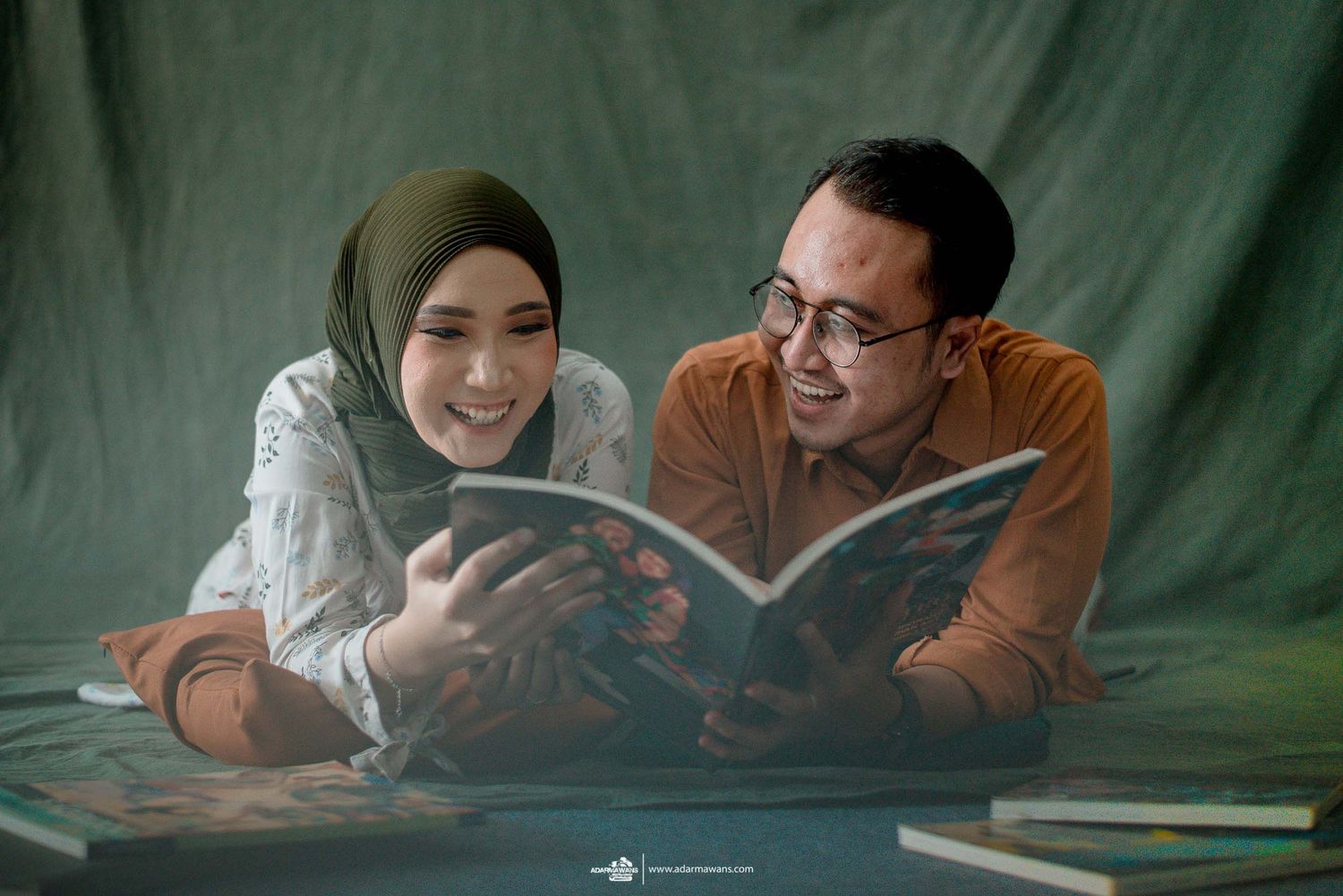 A couple lying down reading a book together against a green backdrop during an intimate photoshoot session.