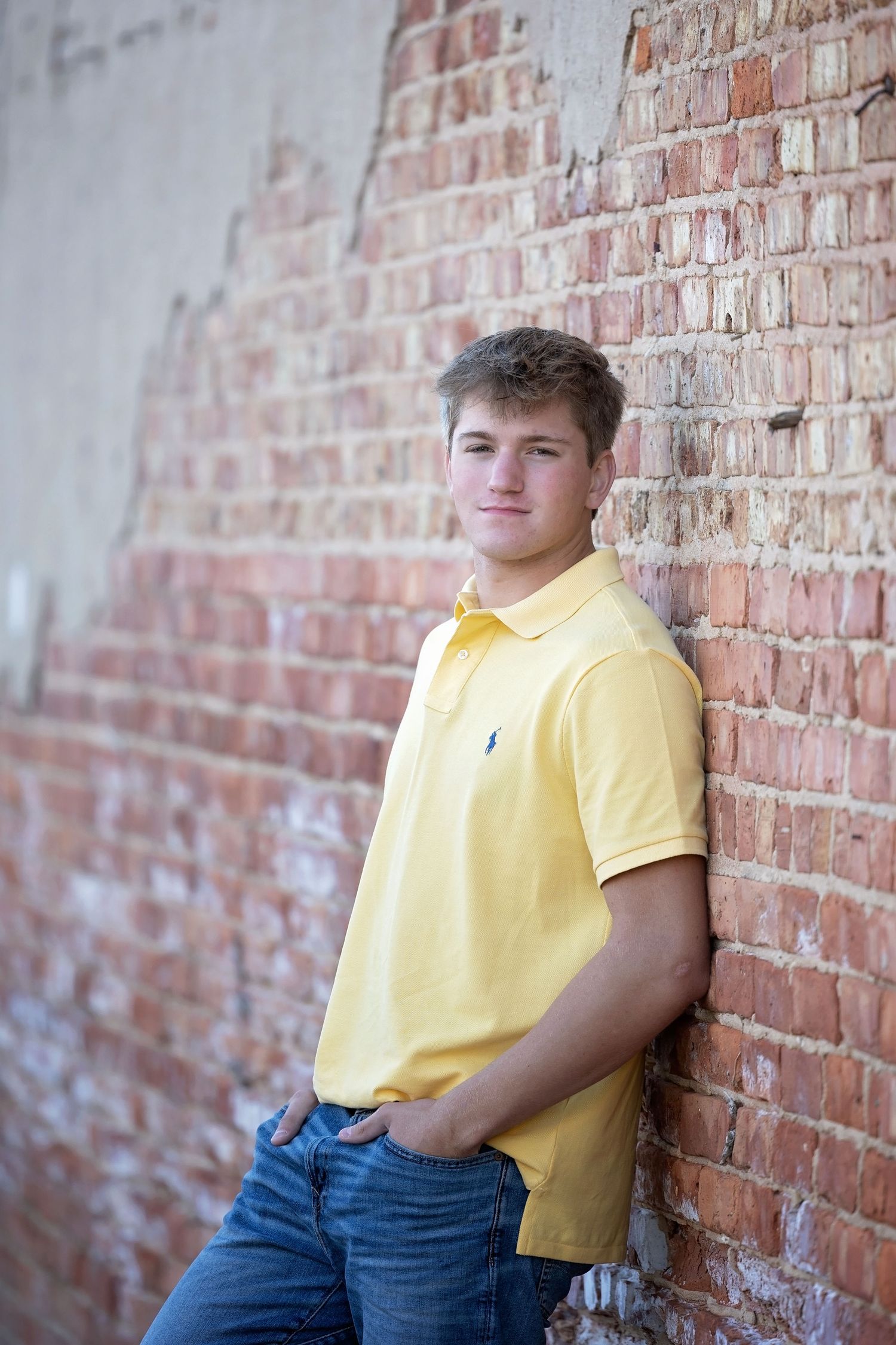 Young person in yellow polo shirt leaning against rustic brick wall in casual outdoor portrait.