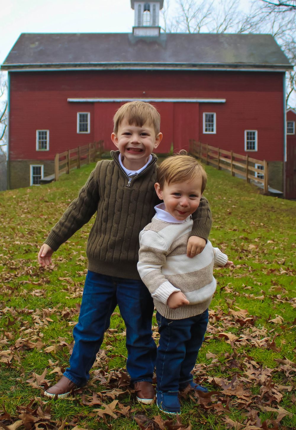 Two children laughing together in front of red barn on autumn day.