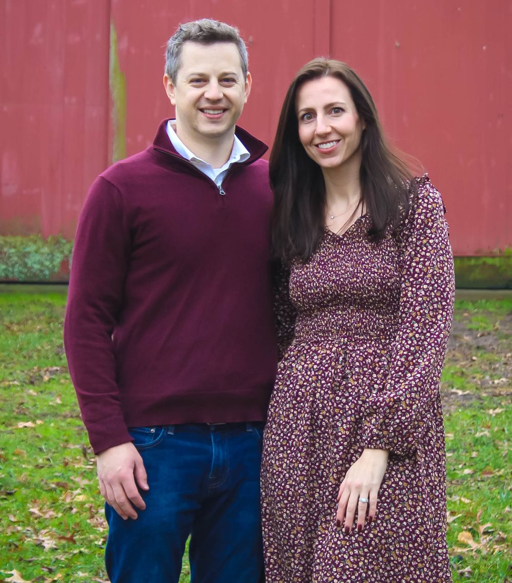 A couple poses outdoors in complementary burgundy attire against a red barn backdrop.