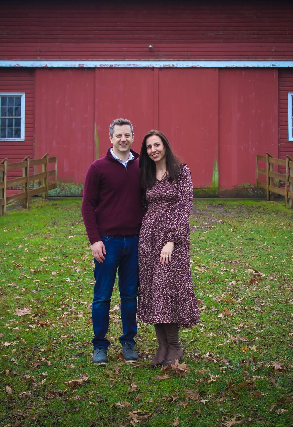 Couple poses together against red barn wall on green grass.