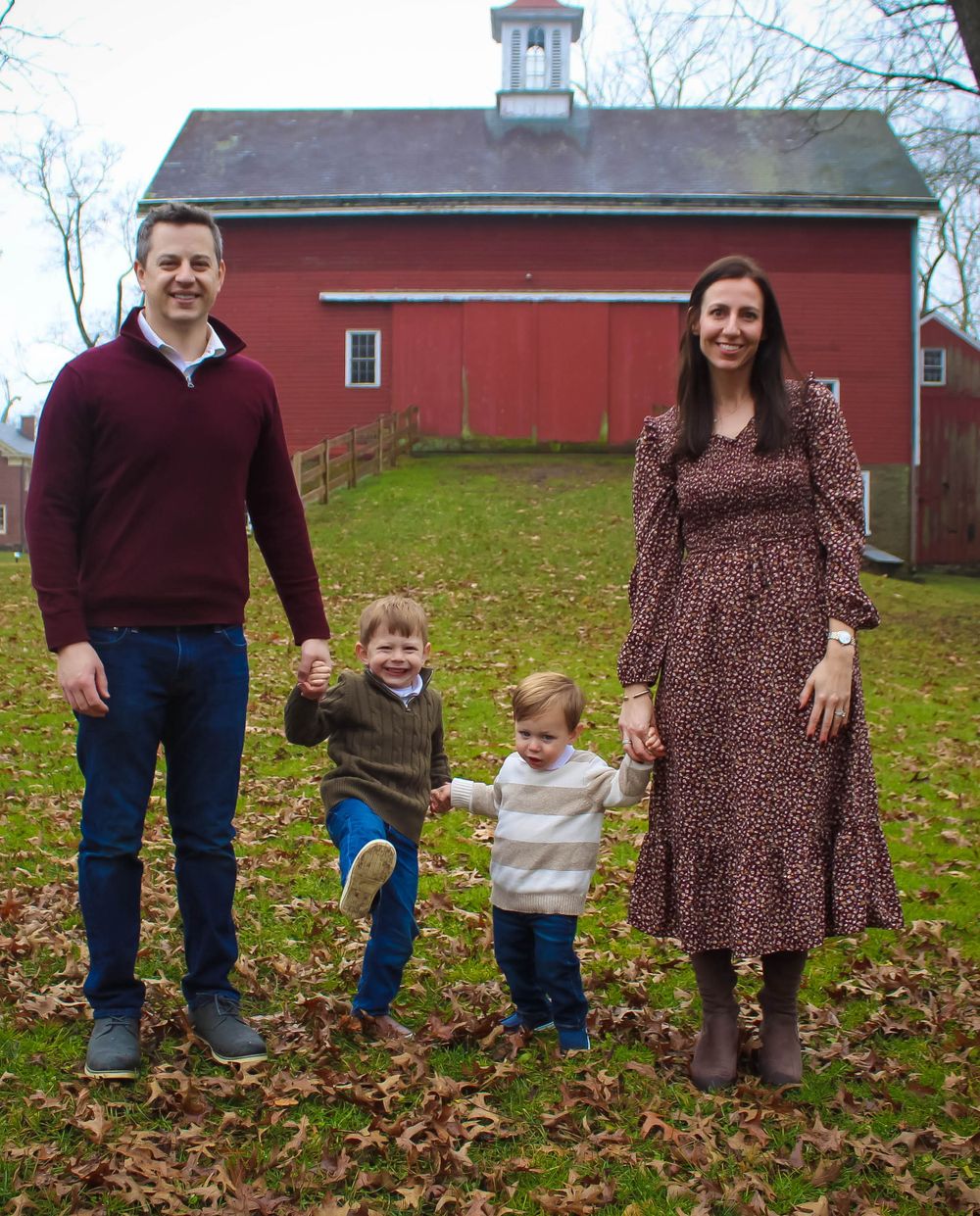 Family holding hands while standing on fallen leaves in front of red barn.