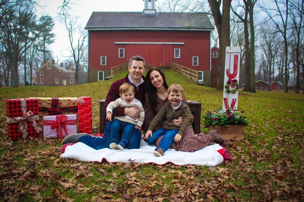 Family poses on blanket with wrapped Christmas presents in front of rustic red barn.