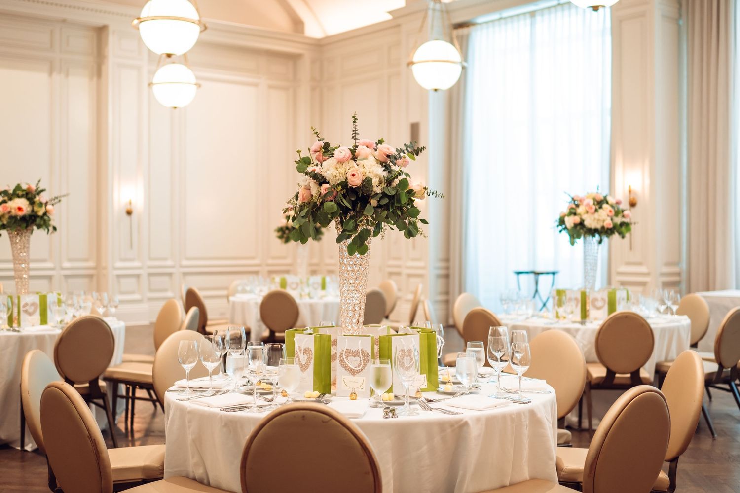 Elegant white wedding reception venue with round tables, floral centerpieces and hanging chandeliers.