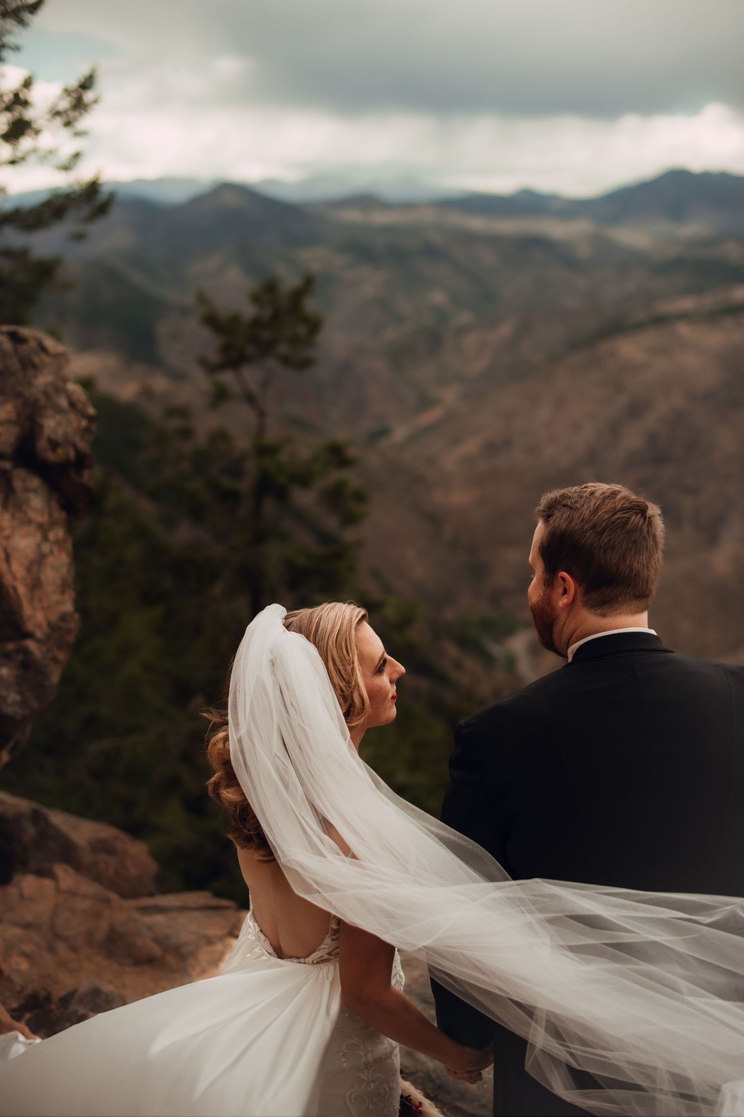 Wedding couple overlooking mountain vista as bride's veil flows in the wind.