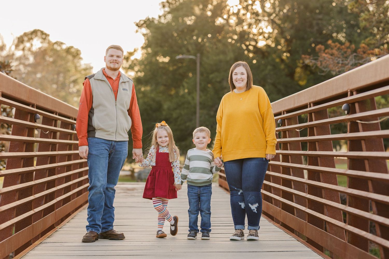 Family of four walking together on wooden bridge during sunset photo session.