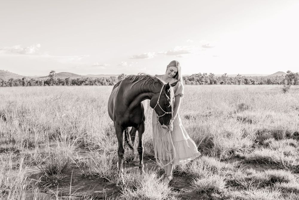 Monochrome portrait of a person in a light dress standing beside a dark horse in a grassy meadow.