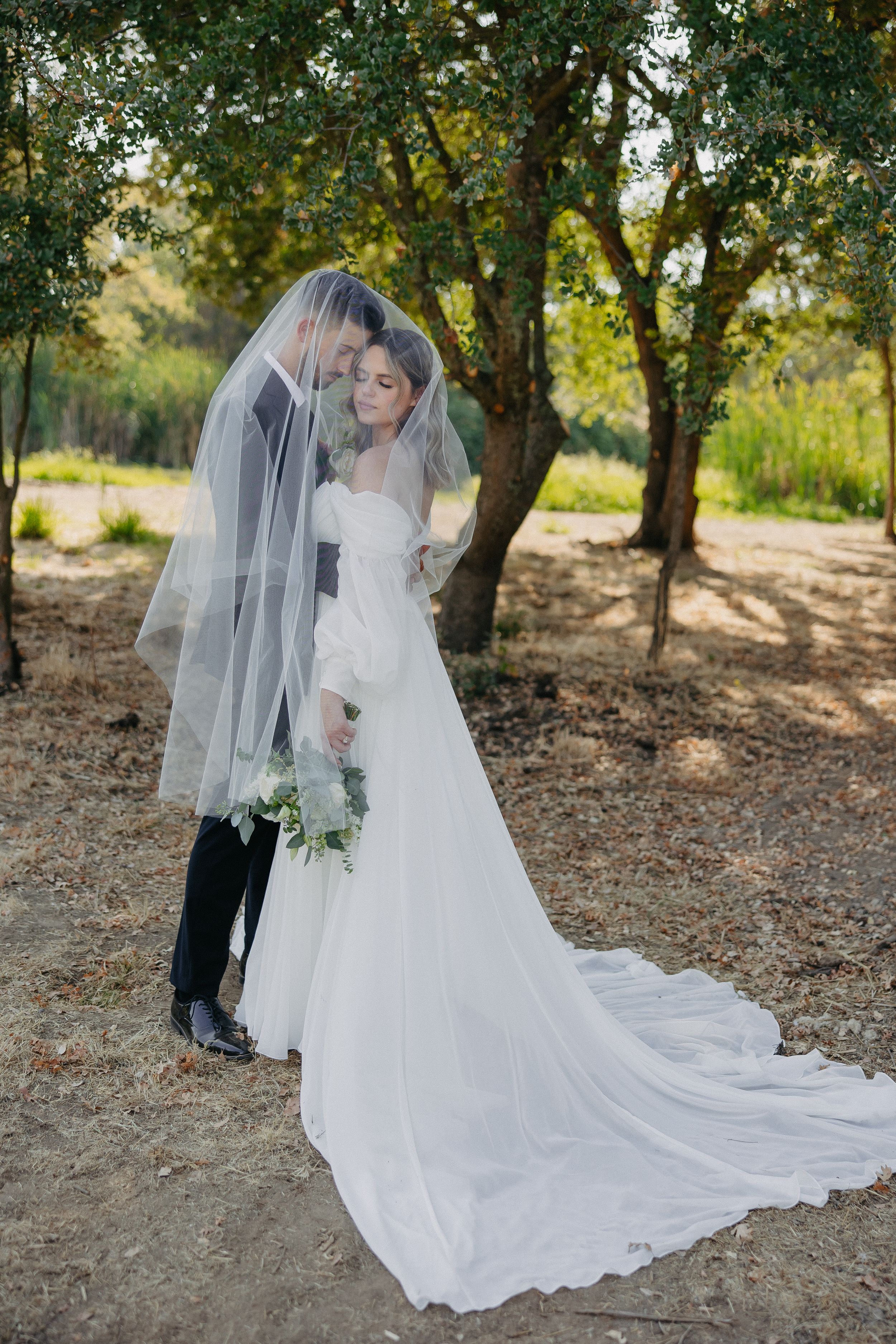 A romantic wedding photo under a tree shows a couple sharing an intimate moment beneath a flowing veil.