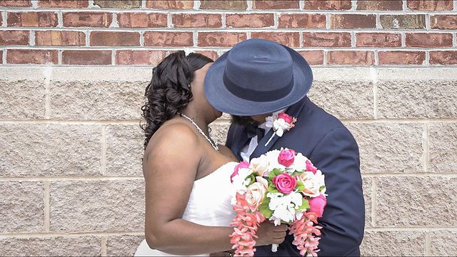 Romantic wedding photo moment with couple and pink floral bouquet against brick wall.