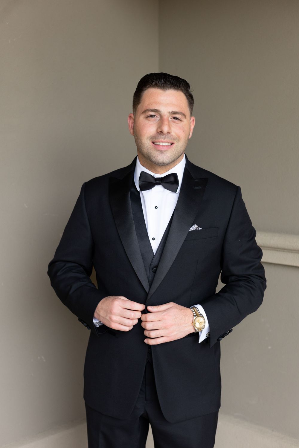 Groom in classic black tuxedo and bow tie adjusts his jacket buttons while getting ready.