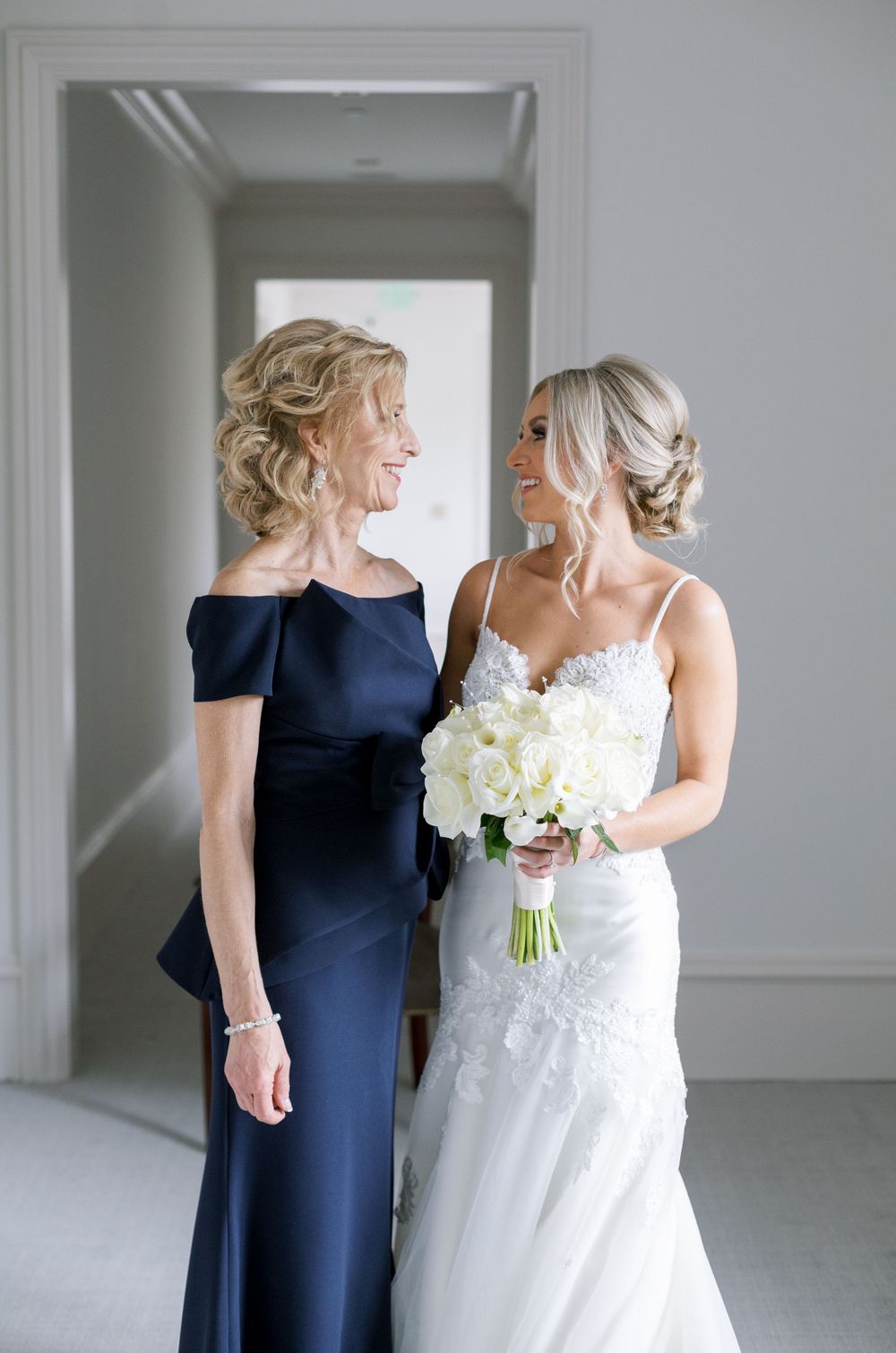 Bride in white lace gown holds white bouquet while sharing moment with woman in navy dress.