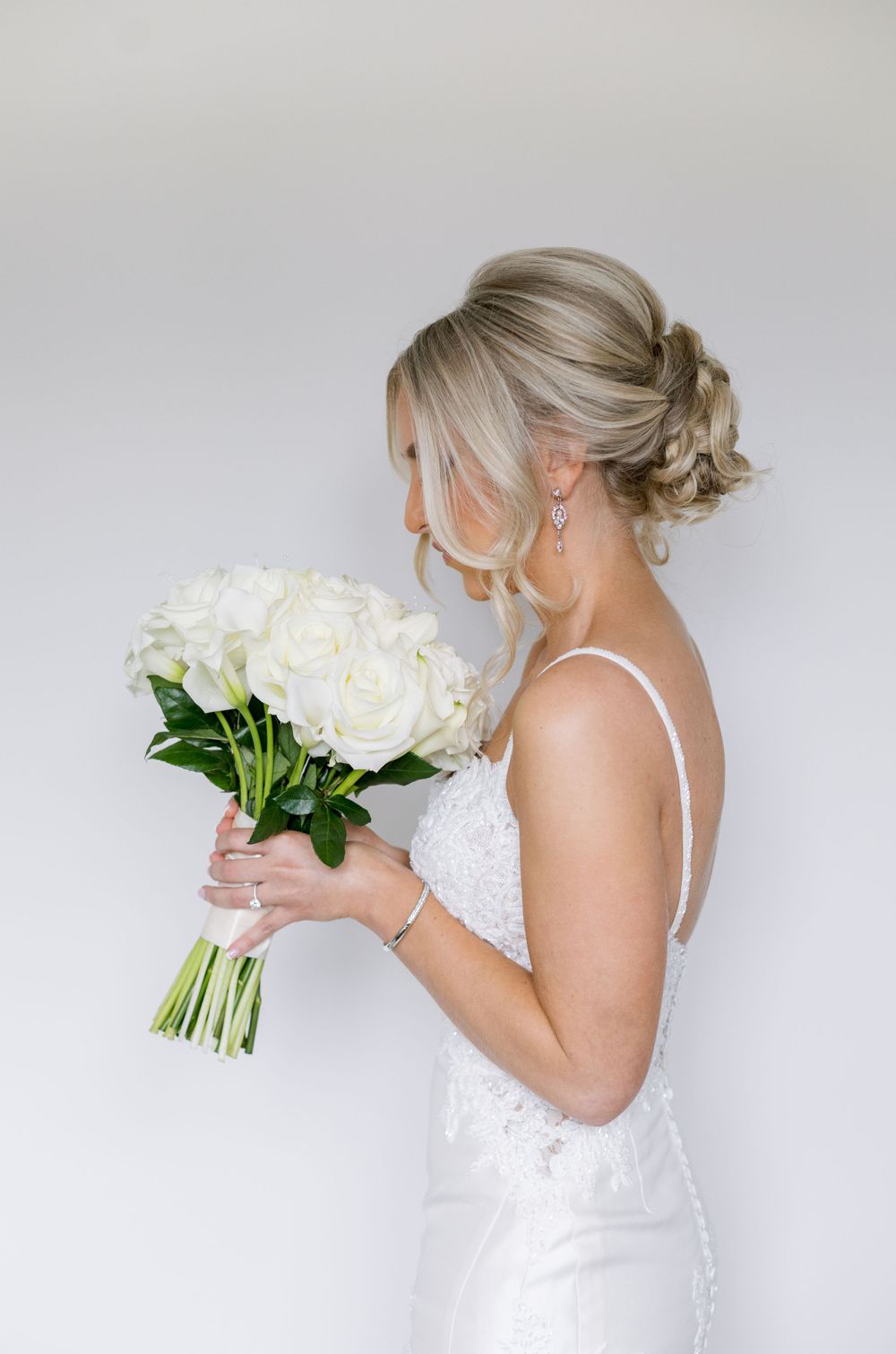 Side profile of bride in white lace gown holding classic white rose bouquet.