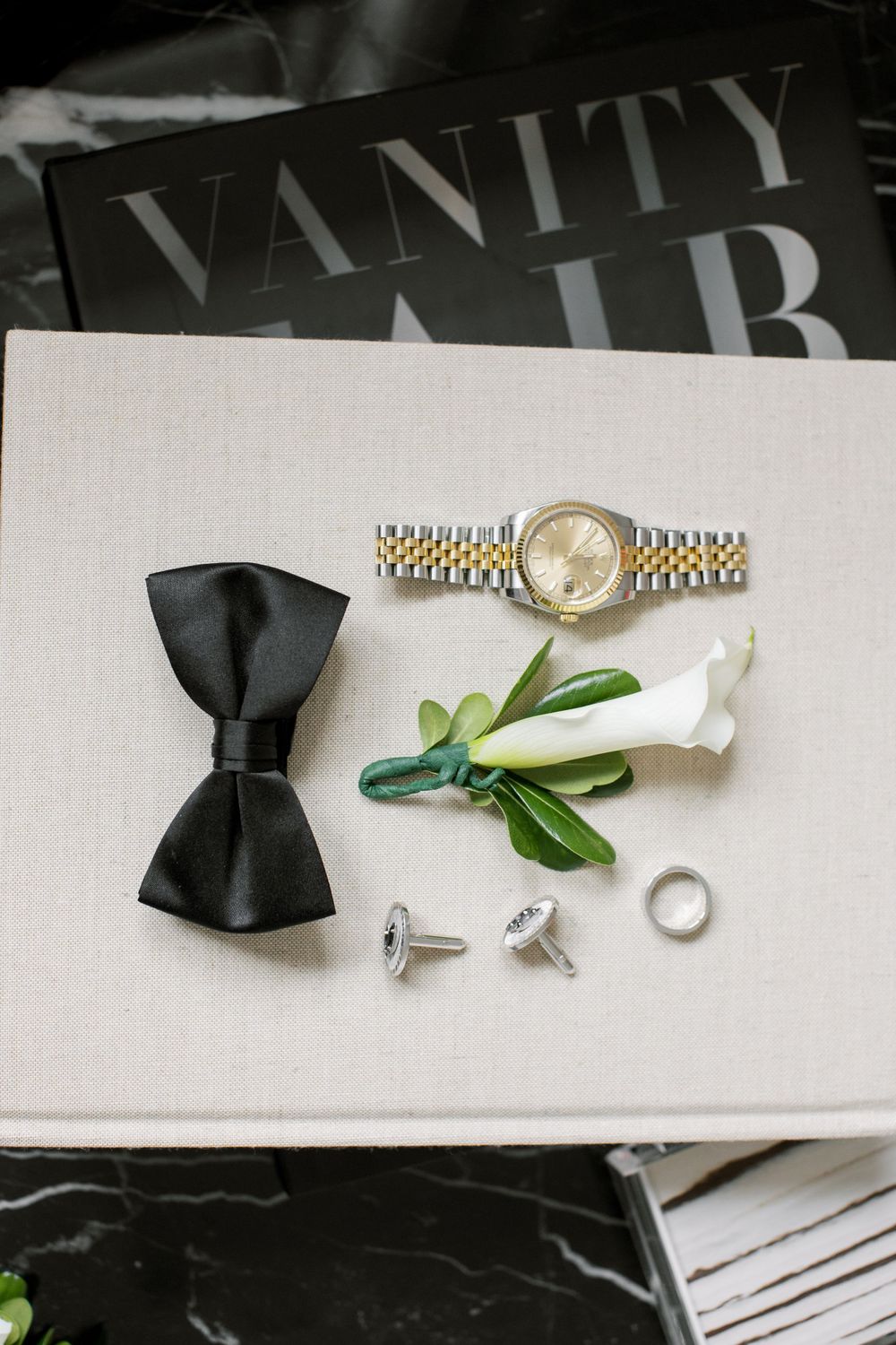 Wedding accessories including black bow tie, watch, boutonniere and rings arranged on white surface.